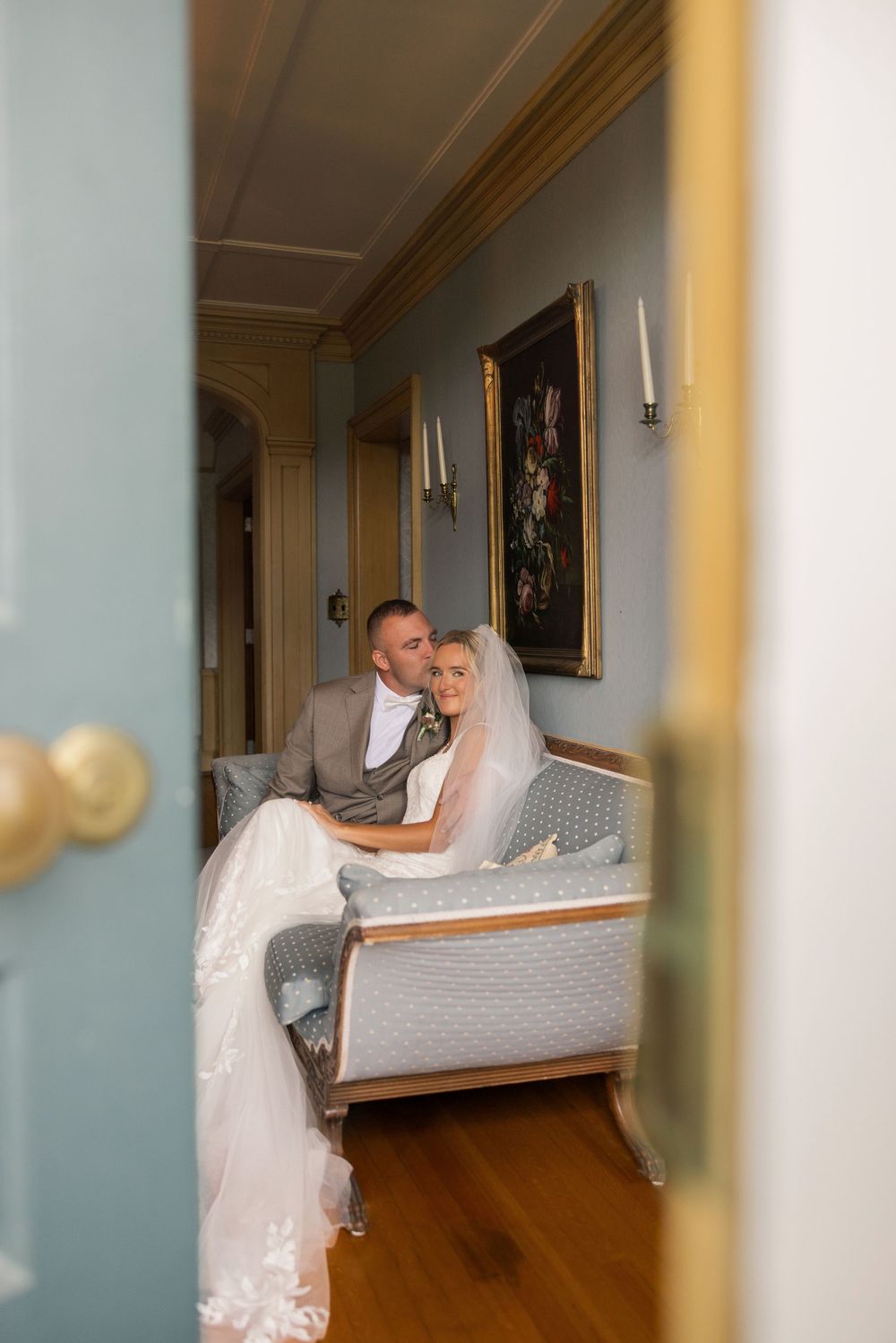 Bride and groom sitting together on an elegant blue settee in a classic decorated room with gold-framed artwork.