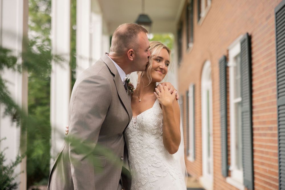 Groom in gray suit kissing bride in white lace wedding dress on cheek outdoors near brick building.