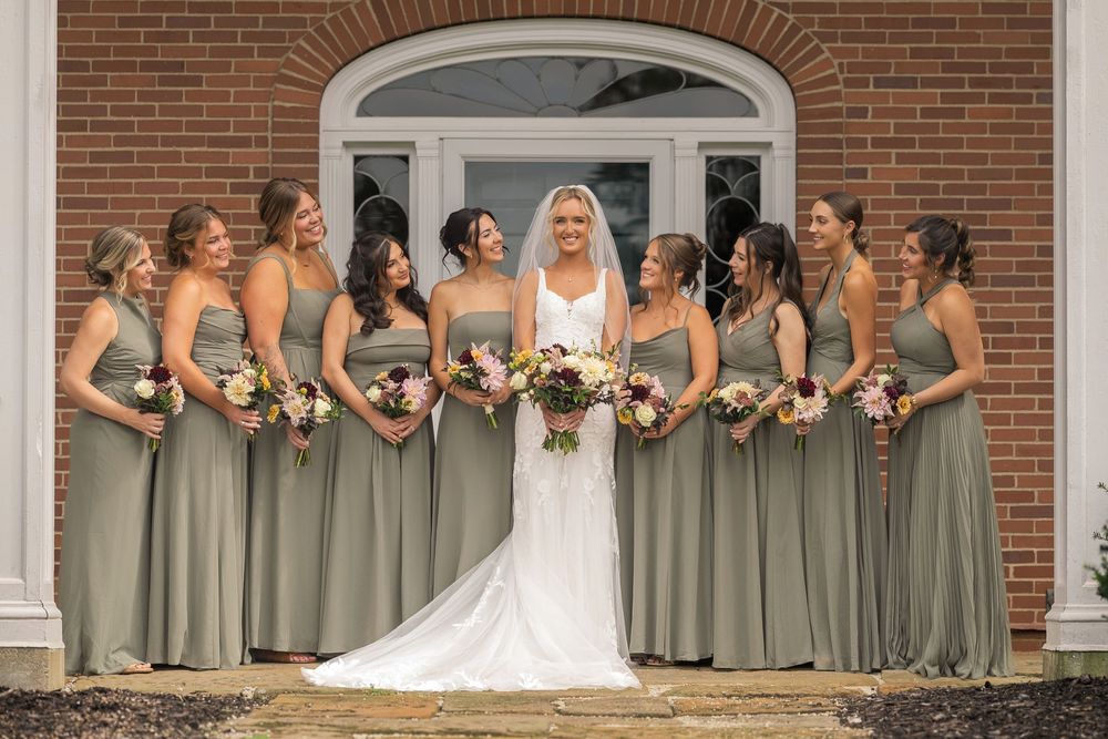 Bride in white gown with bridesmaids in sage green dresses holding bouquets outside brick building.
