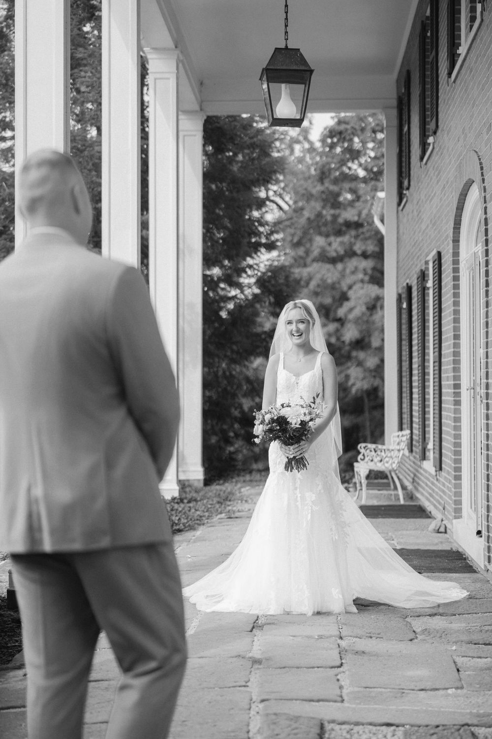 Black and white photo of a bride in a wedding gown holding a bouquet during a first look with her groom on a stone porch.