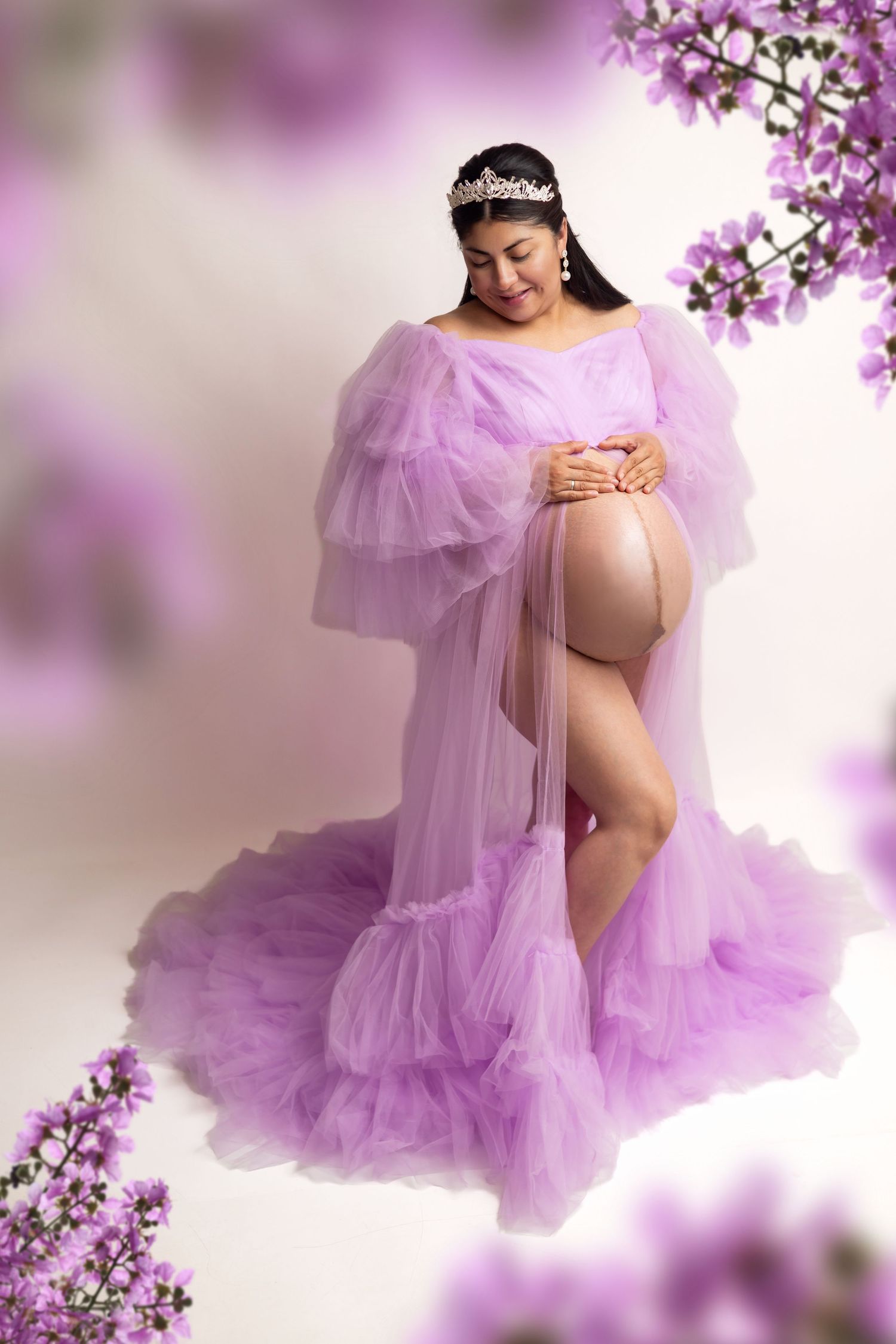 Pregnant woman in flowing purple tulle gown holding baby bump, wearing tiara, surrounded by purple flowers.