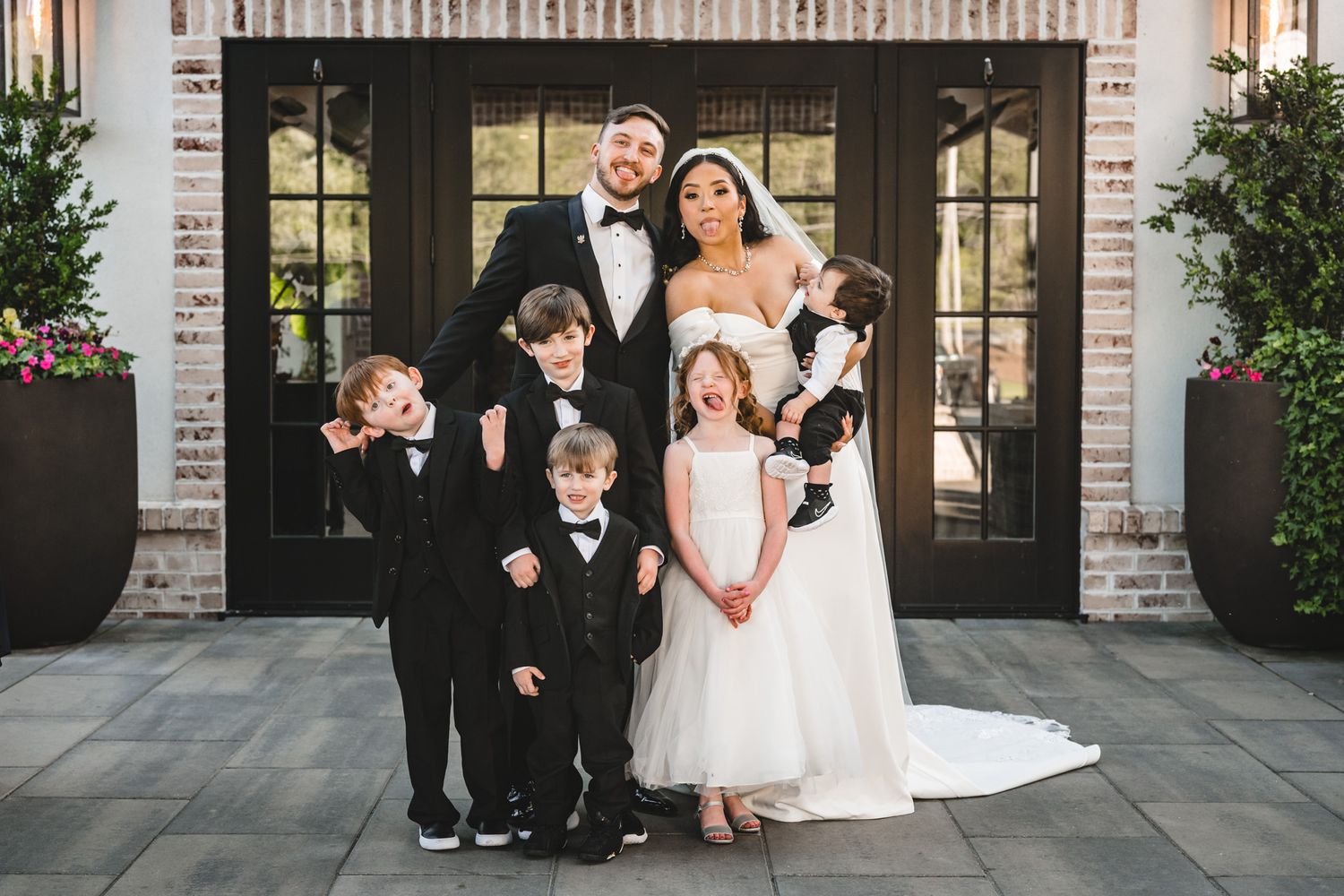 Wedding group photo with bridal party in front of elegant white doors and brick facade.