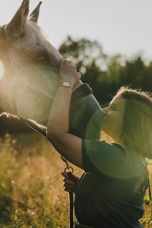 Woman in a green shirt gently kisses the nose of a gray horse in warm, glowing sunset light.