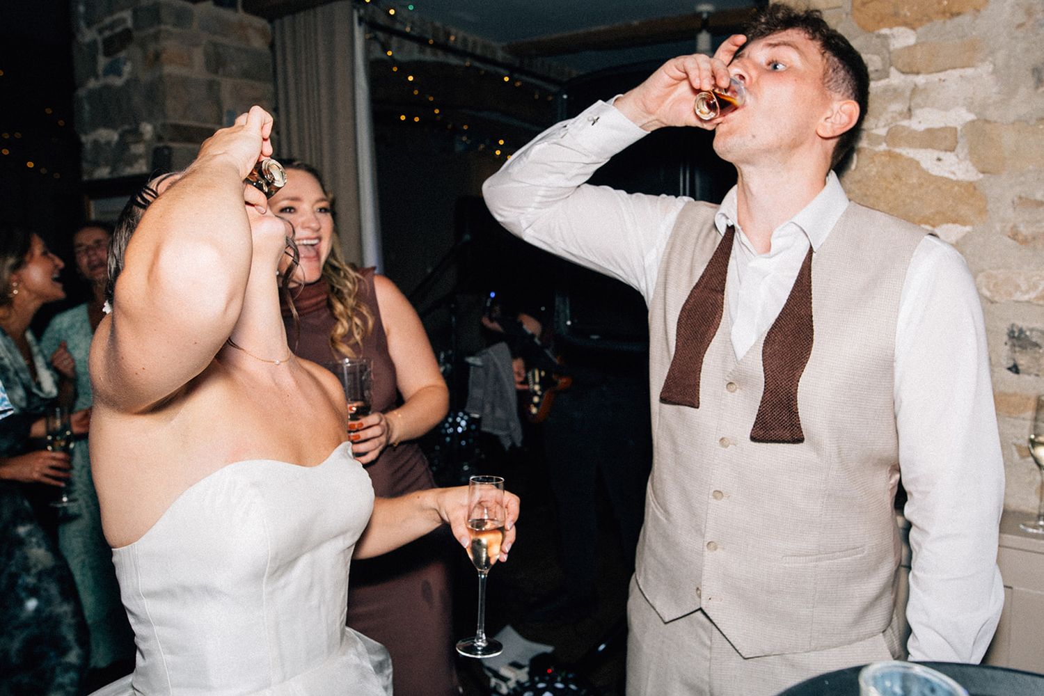 Bride in a white strapless gown and groom in a tan vest and bow tie drink shots together at a lively wedding reception.
