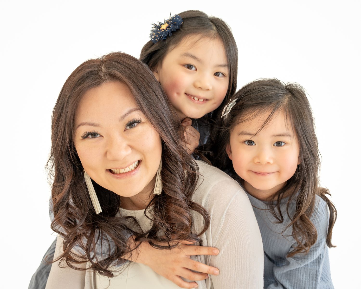 Mother smiling with her two young daughters gathered closely around her during a family portrait against a white background.