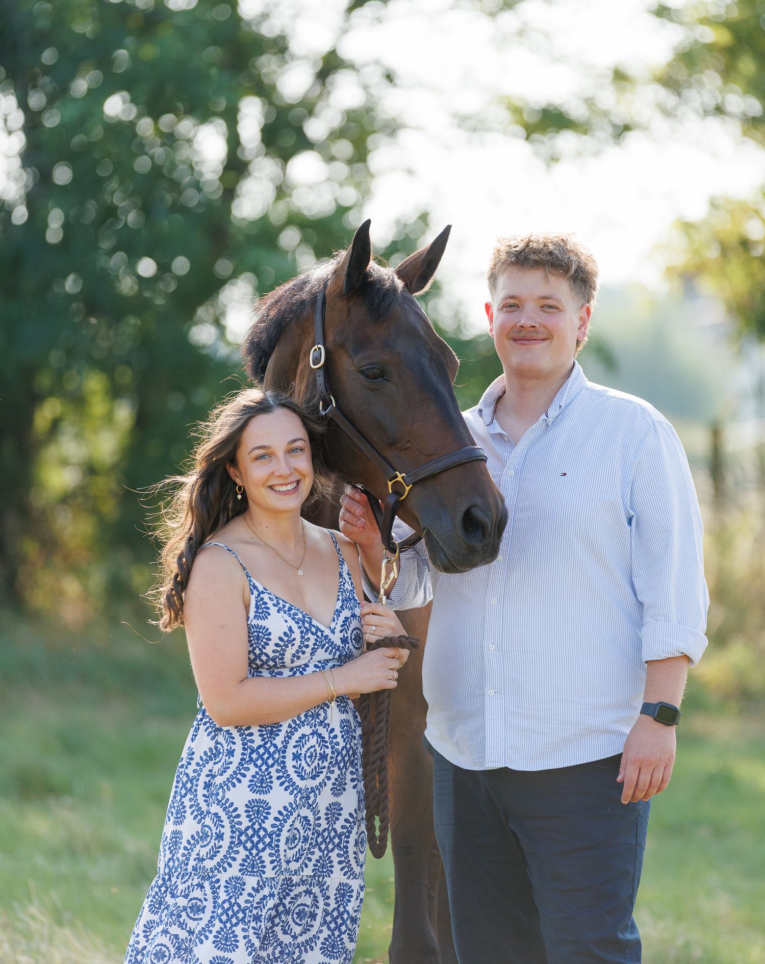 Woman in light blue gown walking with a bay horse on a sunlit tree-lined gravel path.