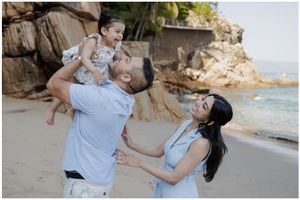 Family of three enjoying a playful moment on a tropical beach, father lifting toddler while mother watches joyfully.