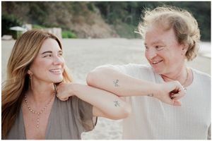 Two people smiling and comparing matching tattoos on their arms at a beach.