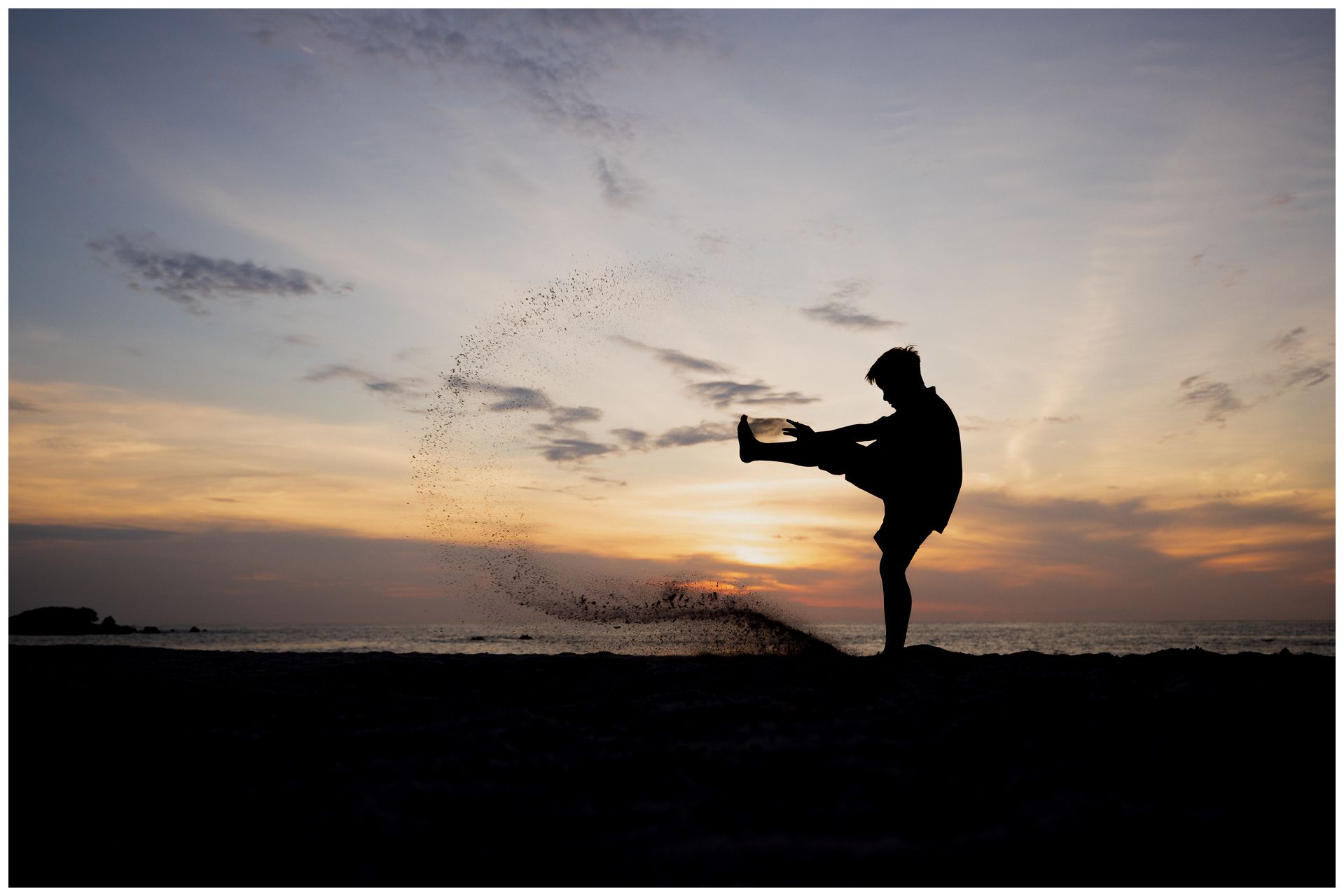 Silhouette of a person performing a high kick at sunset on a beach, with water splashing dramatically.