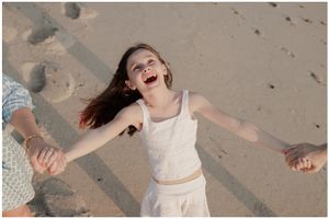 A joyful girl laughs with arms outstretched, held by hands on a sandy beach.
