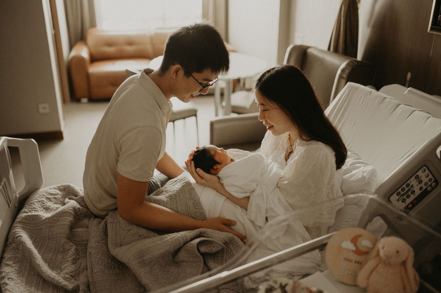 Father and mother in a hospital room lovingly look at their newborn baby, with plush toys resting at the foot of the bed.