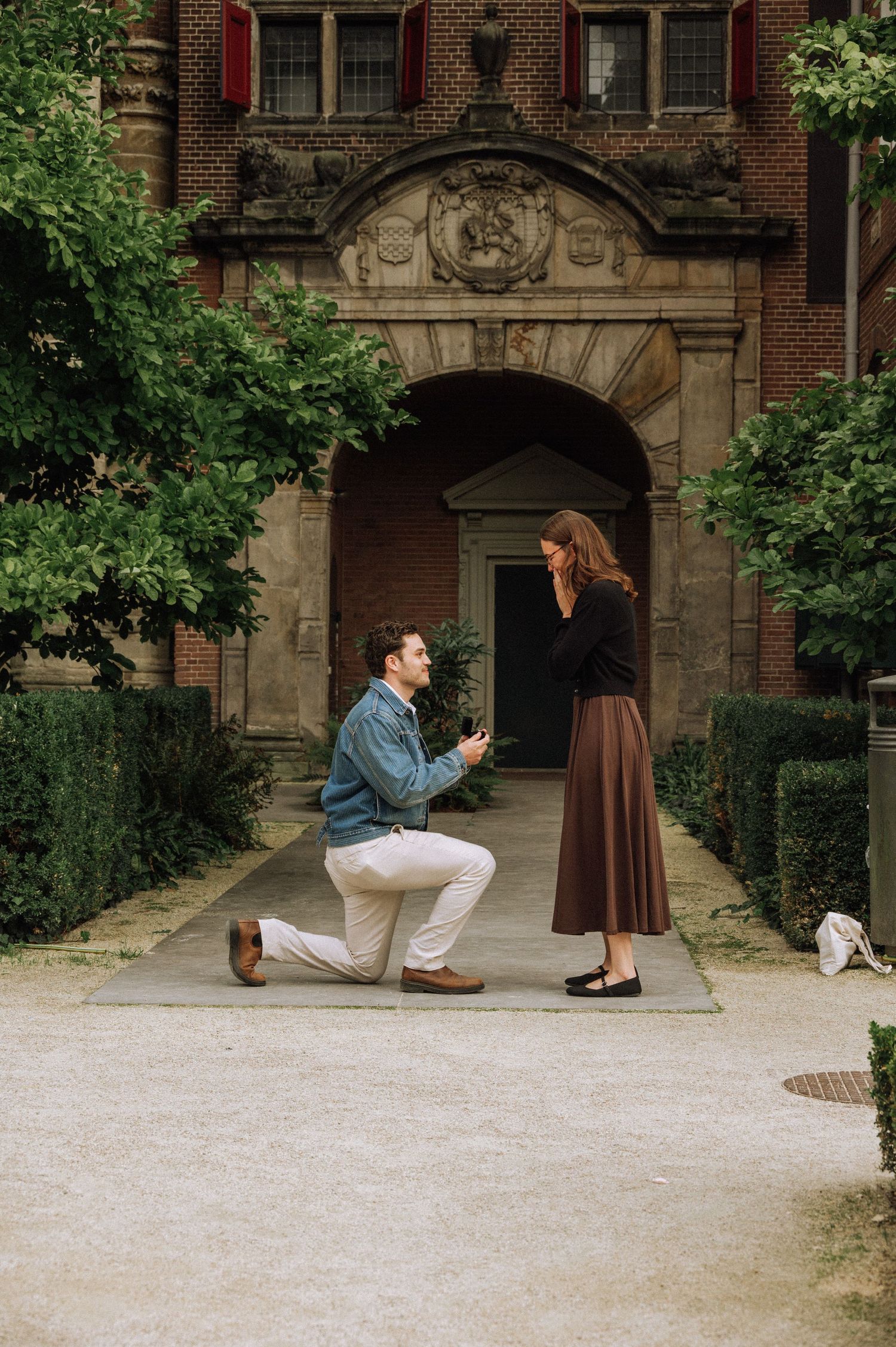 Man in a blue jacket kneels on one knee proposing to a woman in a long brown skirt outside a historic brick building archway. Rijksmuseum
