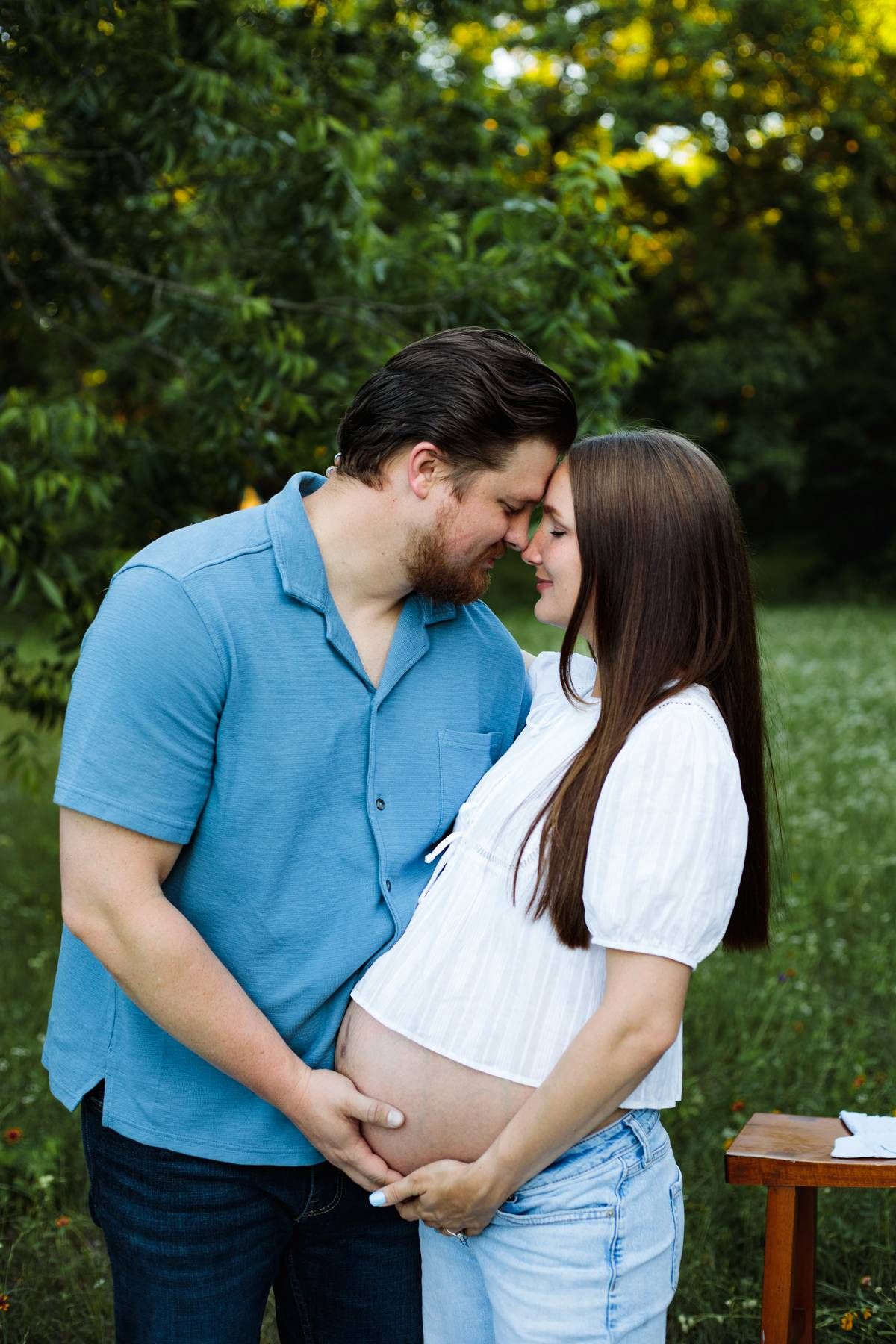 Bearded man in a blue polo and pregnant woman in a white blouse touch foreheads and cradle her baby bump outdoors.