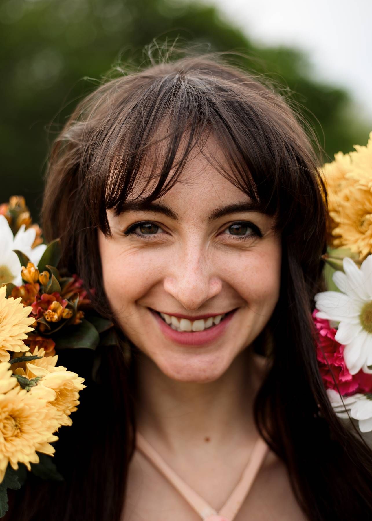 A smiling woman with long dark hair and bangs poses outdoors surrounded by yellow, white and pink flowers.