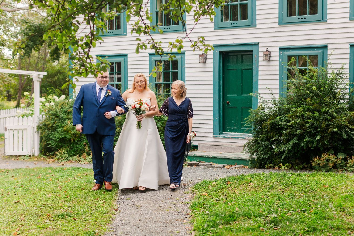 Bride in a white gown walks arm in arm with a man in a navy suit and a woman in a navy dress past a white clapboard house with green doors.