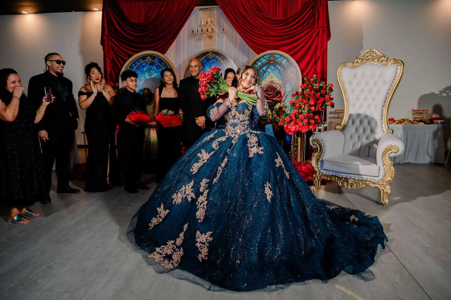 Young woman in a sparkling navy blue quinceañera ball gown holding a bouquet, posed with guests in black beside a white throne chair.