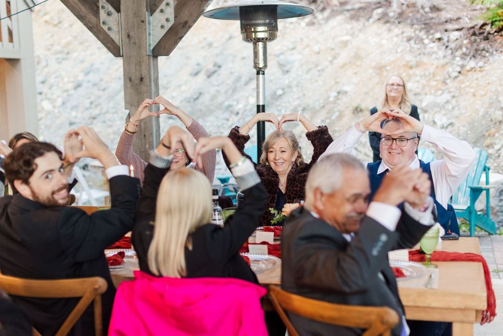 Wedding guests seated at a long outdoor table raise their joined hands in a toast under a patio heater with a rocky hillside behind them.