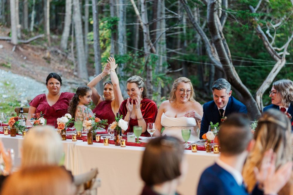 Smiling wedding guests in red and white outfits sit and toast at a long reception table with flowers, set outdoors among tall forest trees.