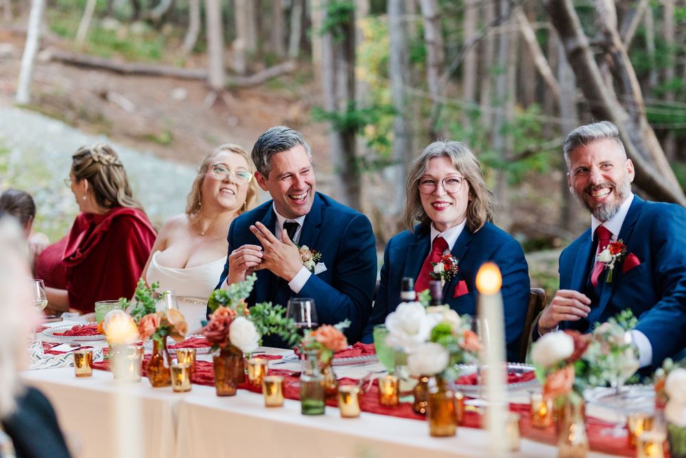 Four smiling guests in navy suits and burgundy attire sit at a candlelit wedding table with floral centerpieces in a forest setting.