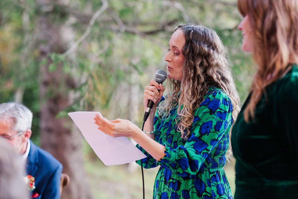A woman with long curly hair in a green patterned dress reads from a paper into a microphone during an outdoor ceremony among trees.