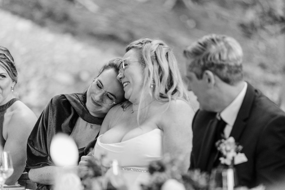 Black and white photo of two women laughing and embracing at a wedding reception while a man in a suit sits beside them.