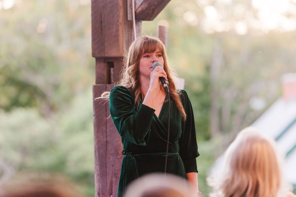 Woman with long hair and bangs in a green velvet dress sings into a microphone beside a wooden post at an outdoor event.
