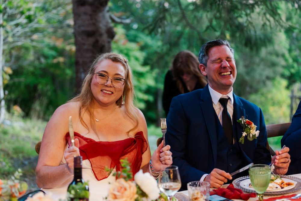 Smiling woman in a red dress and laughing man in a navy suit with a floral boutonniere sit at an outdoor wedding reception table.