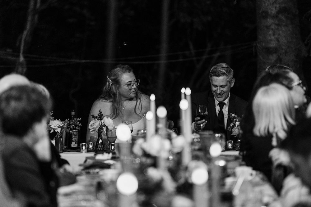 Black and white photo of a smiling couple seated at a candlelit wedding reception table surrounded by guests and flowers.