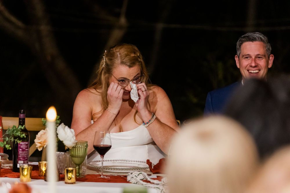 Emotional bride in a white dress wipes happy tears at a candlelit wedding reception table while a smiling man in a suit looks on.