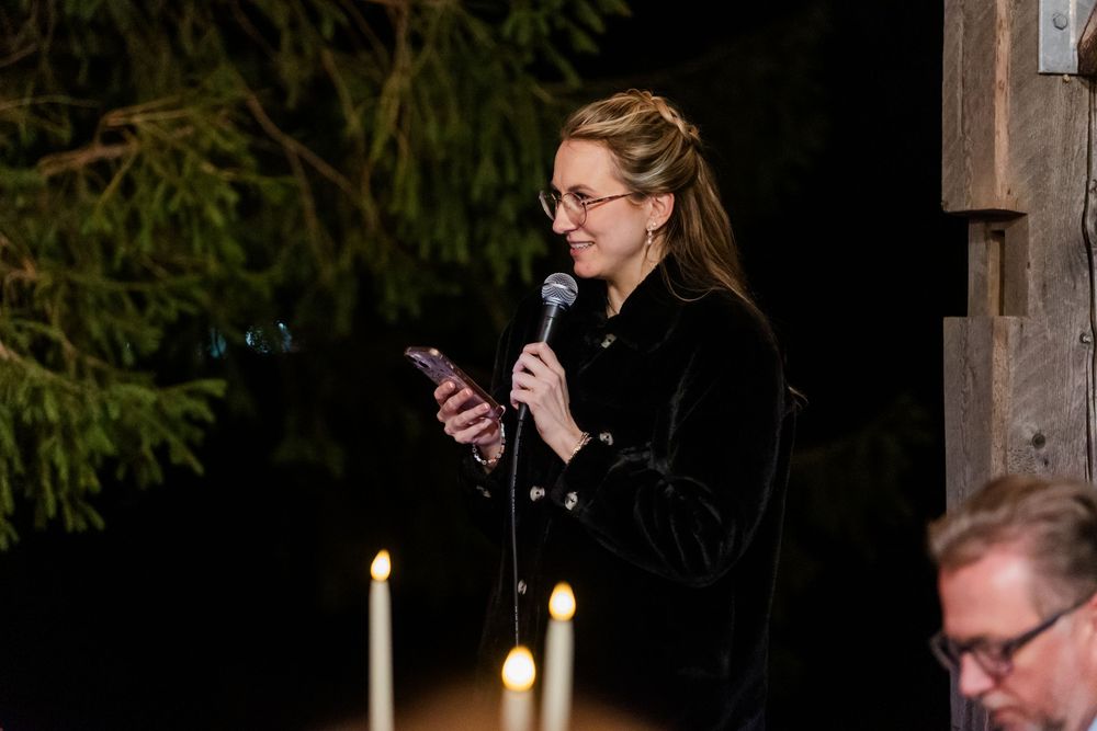 Woman in a black jacket speaks into a microphone while reading from her phone at an evening event, with candles glowing in the foreground.