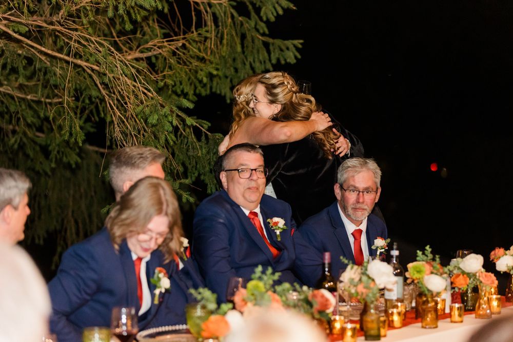 A woman in a brown dress embraces a smiling speaker at a wedding reception while guests in navy suits watch from a decorated table.