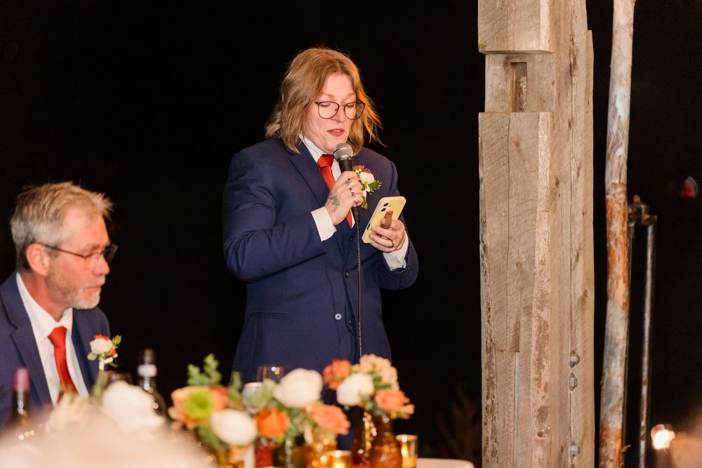 Wedding guest in a navy suit and red tie reads from a phone while giving a toast beside a seated man and a floral centerpiece.