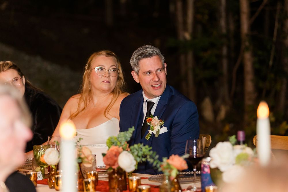 Smiling couple in wedding attire sits at a candlelit reception table decorated with colorful flowers and glowing candles.