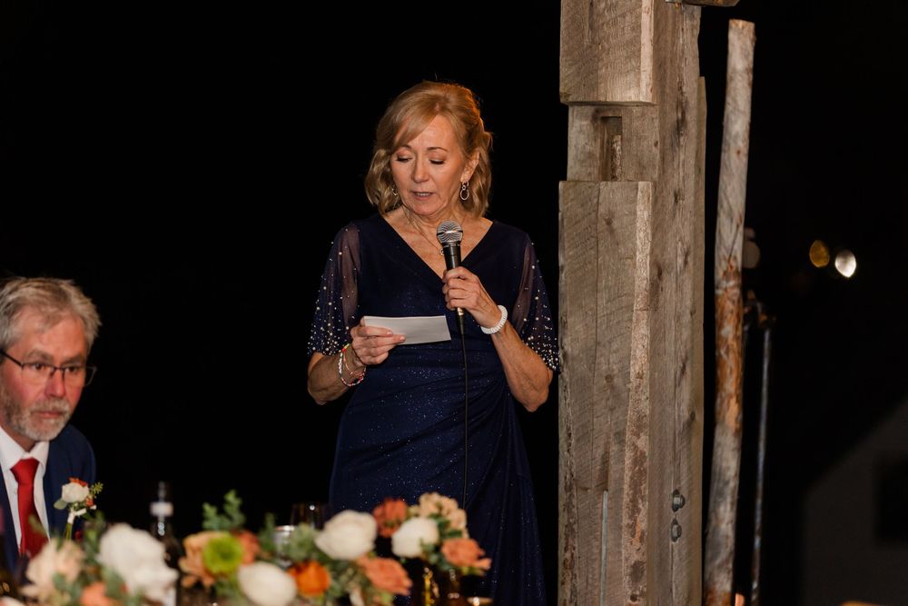 A woman in a navy dress reads from a paper into a microphone while giving a speech at a candlelit reception.