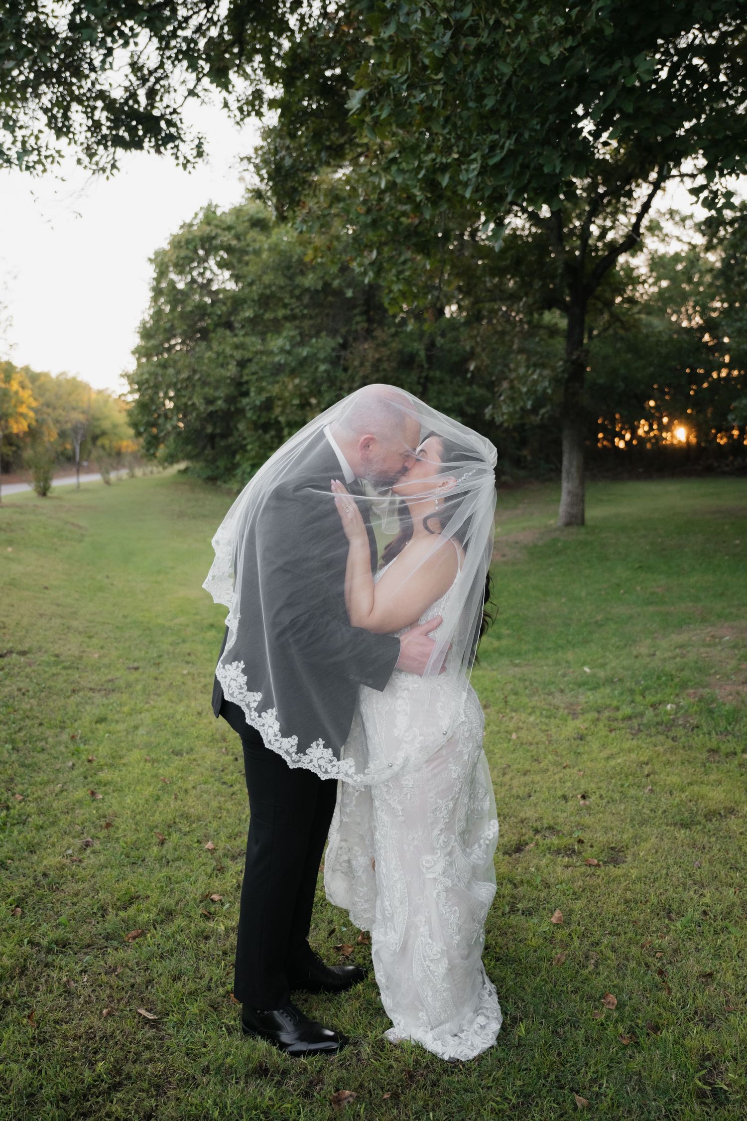 Bride and groom share a kiss beneath a lace-trimmed wedding veil on a green lawn with trees glowing in the sunset behind them.