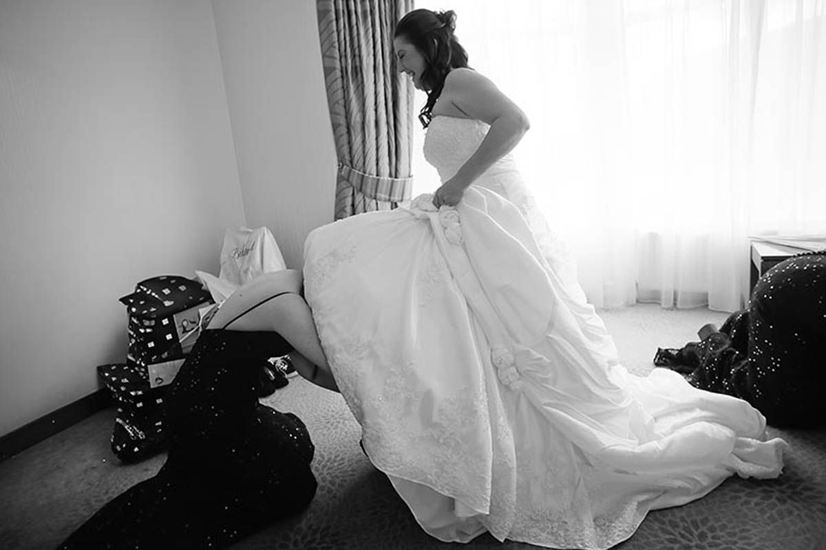 Black and white photo of a bride in a white wedding gown being helped with her dress in a hotel room.