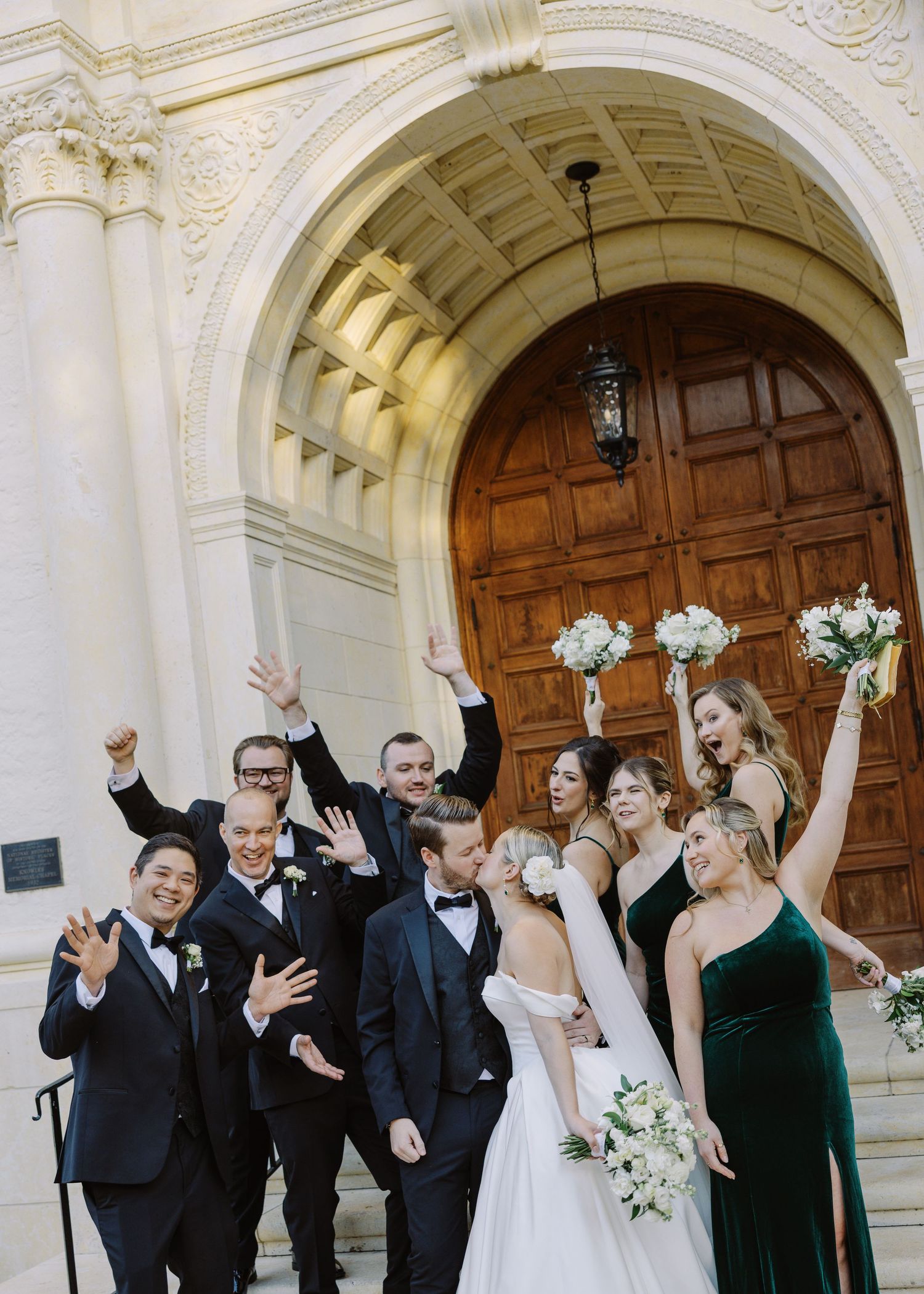 Wedding party celebrating on church steps, bridesmaids in green velvet gowns and groomsmen in black tuxedos cheering joyfully.