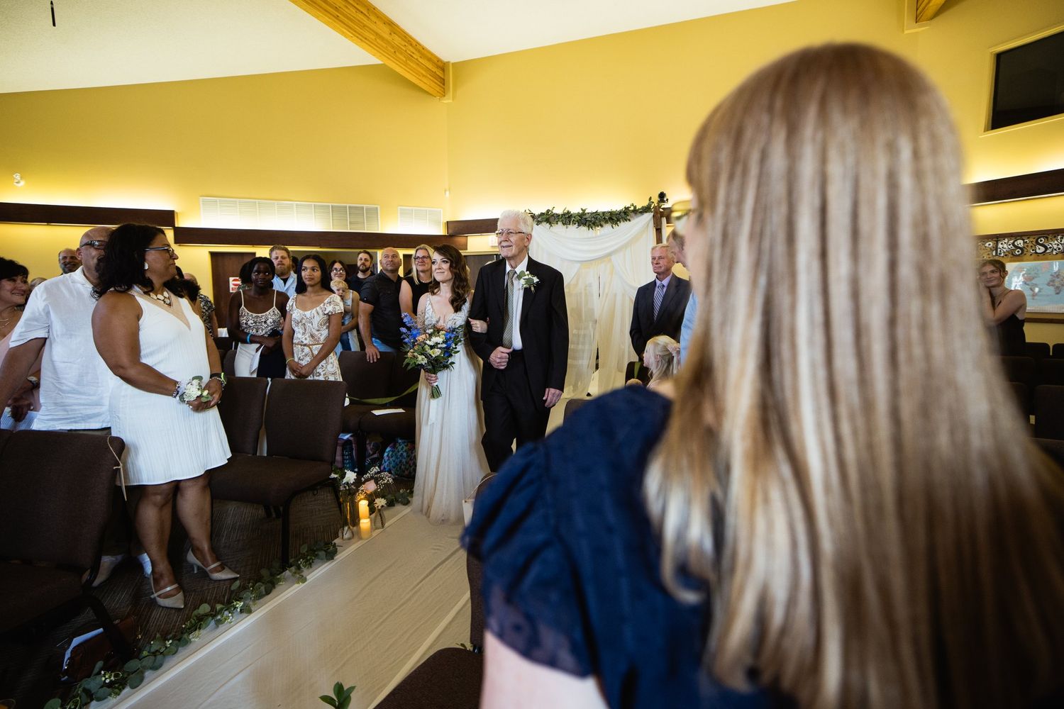 Bride walking down the aisle with her father at King's Gate Church in Cochrane, seen past a guest