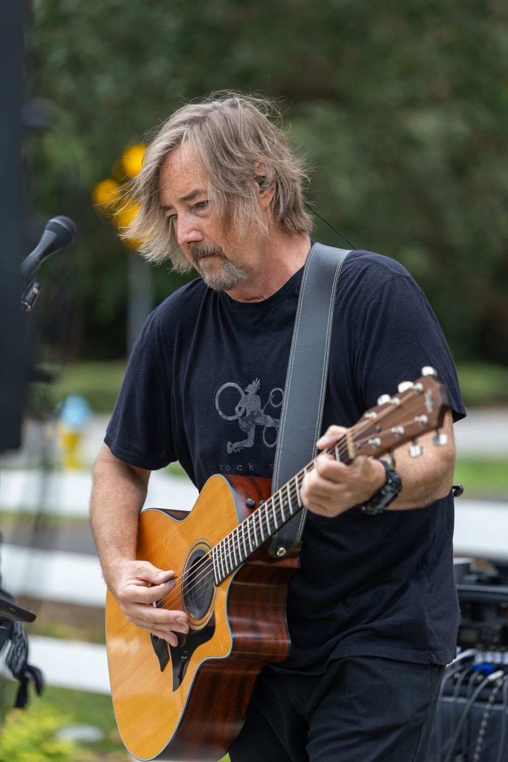 Middle-aged man with gray hair playing acoustic guitar outdoors at a live music performance wearing a black t-shirt.