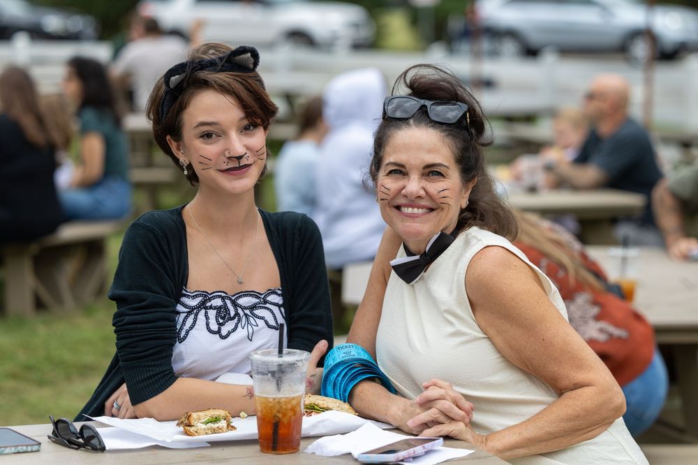 Two women smile at an outdoor dining table with food and drinks at a casual event.