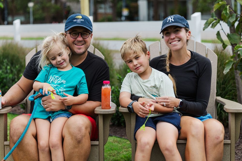 Happy family of four sitting on wooden chairs outdoors, parents wearing NY caps with two young children smiling.