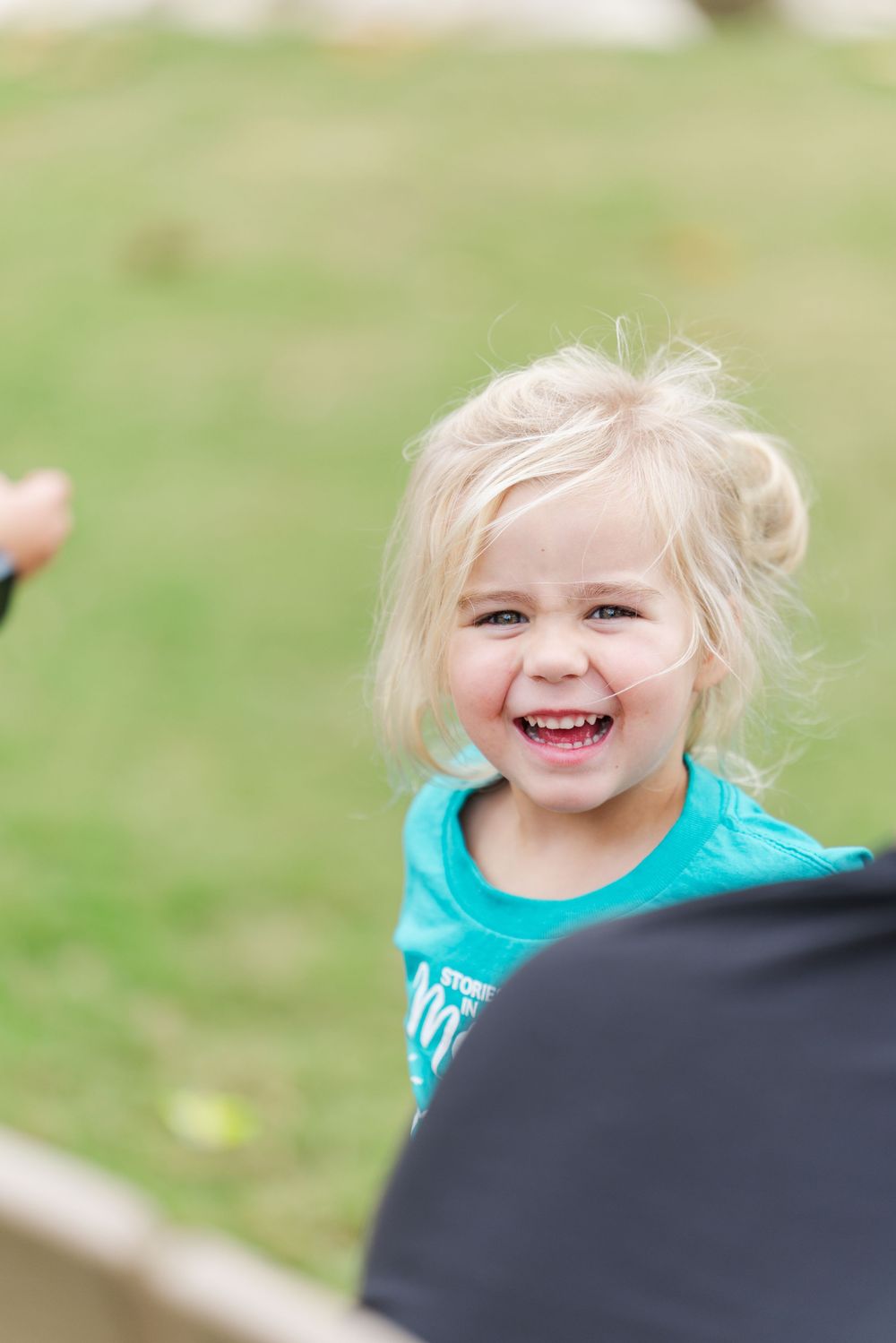 Happy blonde toddler girl in teal shirt laughing outdoors on green grass background.
