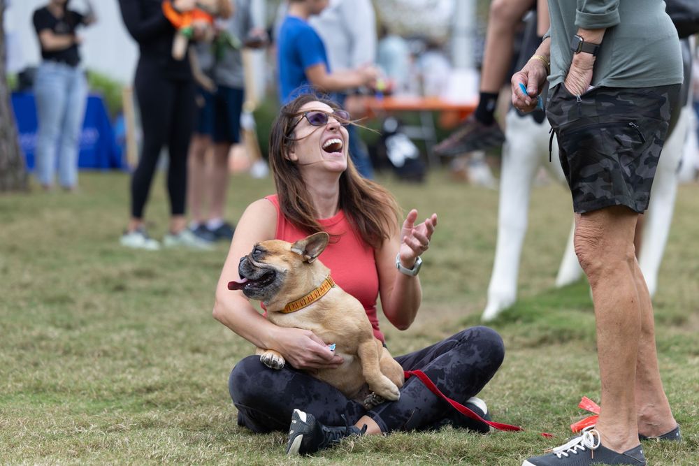 Woman laughing while holding a small dog on grass at an outdoor event with people in the background.