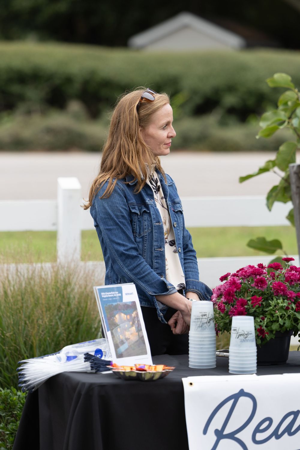 Woman in denim jacket standing at outdoor event table with flowers, brochures, and cups on black tablecloth.