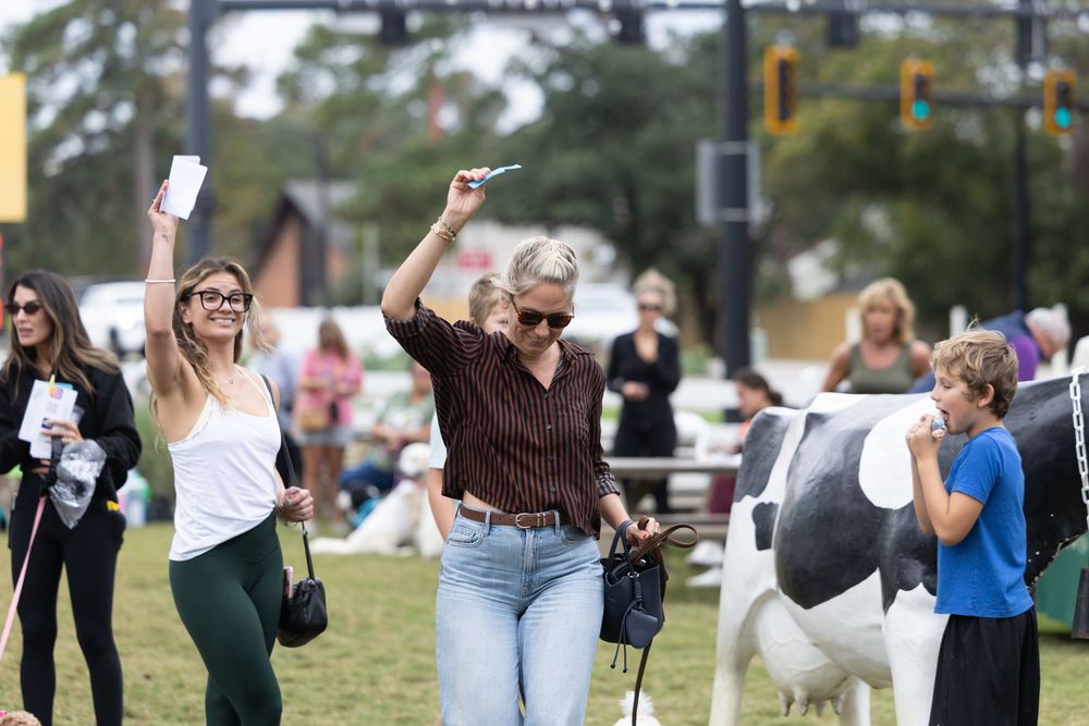 Women walking at an outdoor fair with a cow statue and trees in the background.
