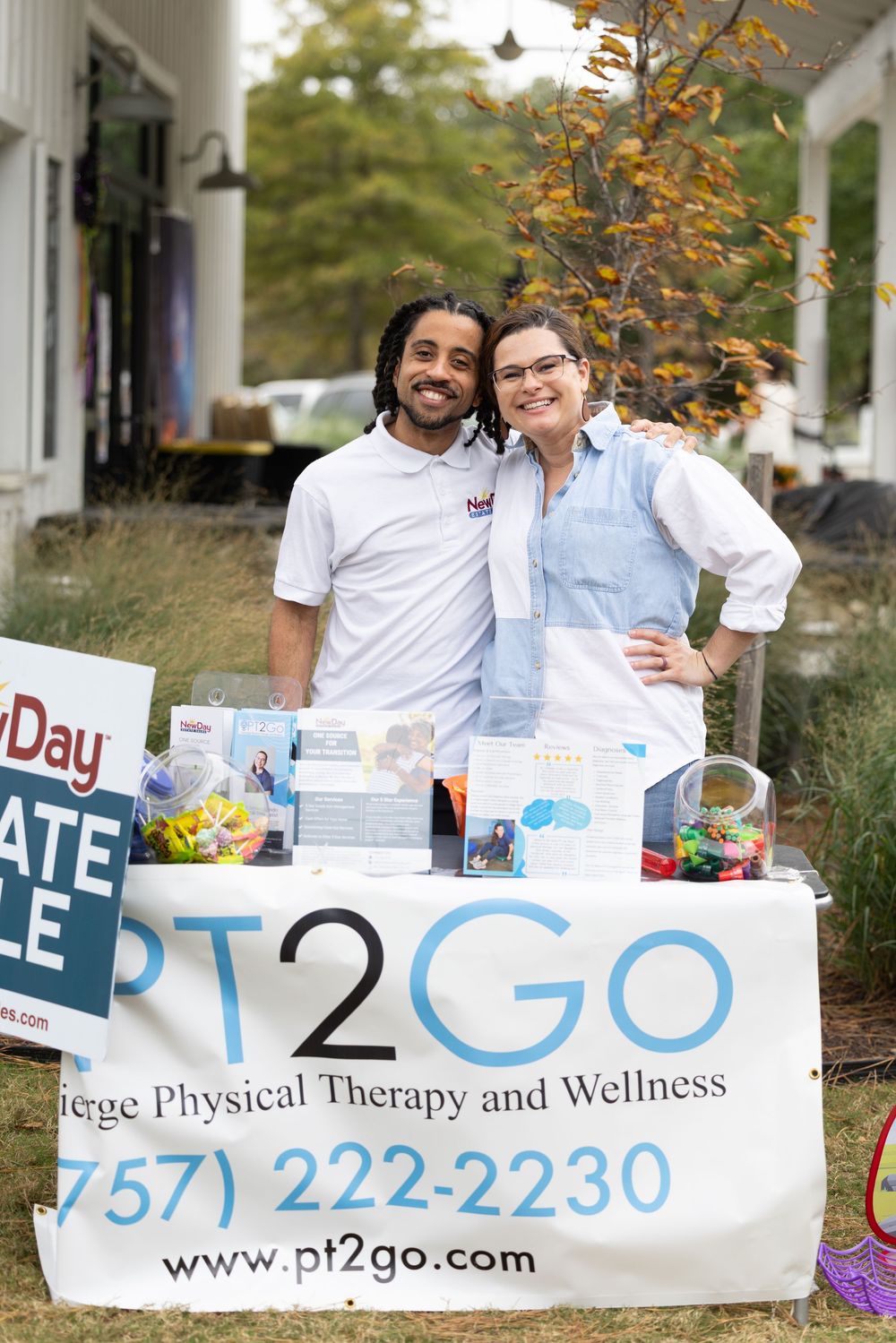 Two smiling staff members pose at a PT2GO Concierge Physical Therapy and Wellness booth at an outdoor event.
