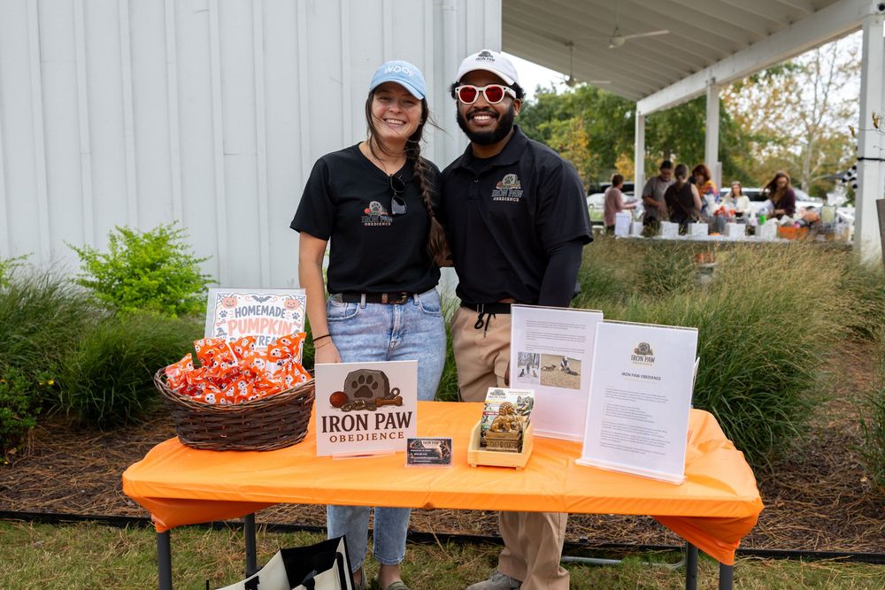 Two Iron Paw Biscuits vendors smile at an outdoor market booth with dog treats and signage on display.