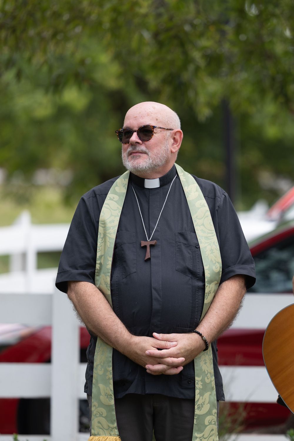Bald priest wearing black clergy shirt, green stole, and cross necklace stands outdoors with sunglasses.