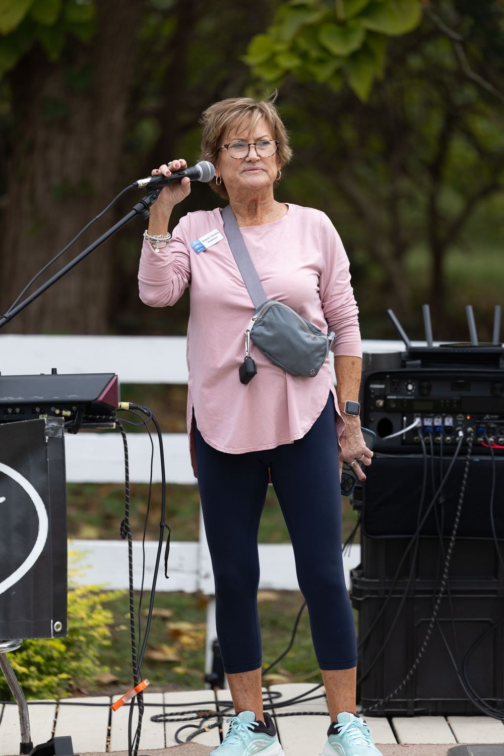 Older woman in pink top and leggings speaks into a microphone at an outdoor event stage.