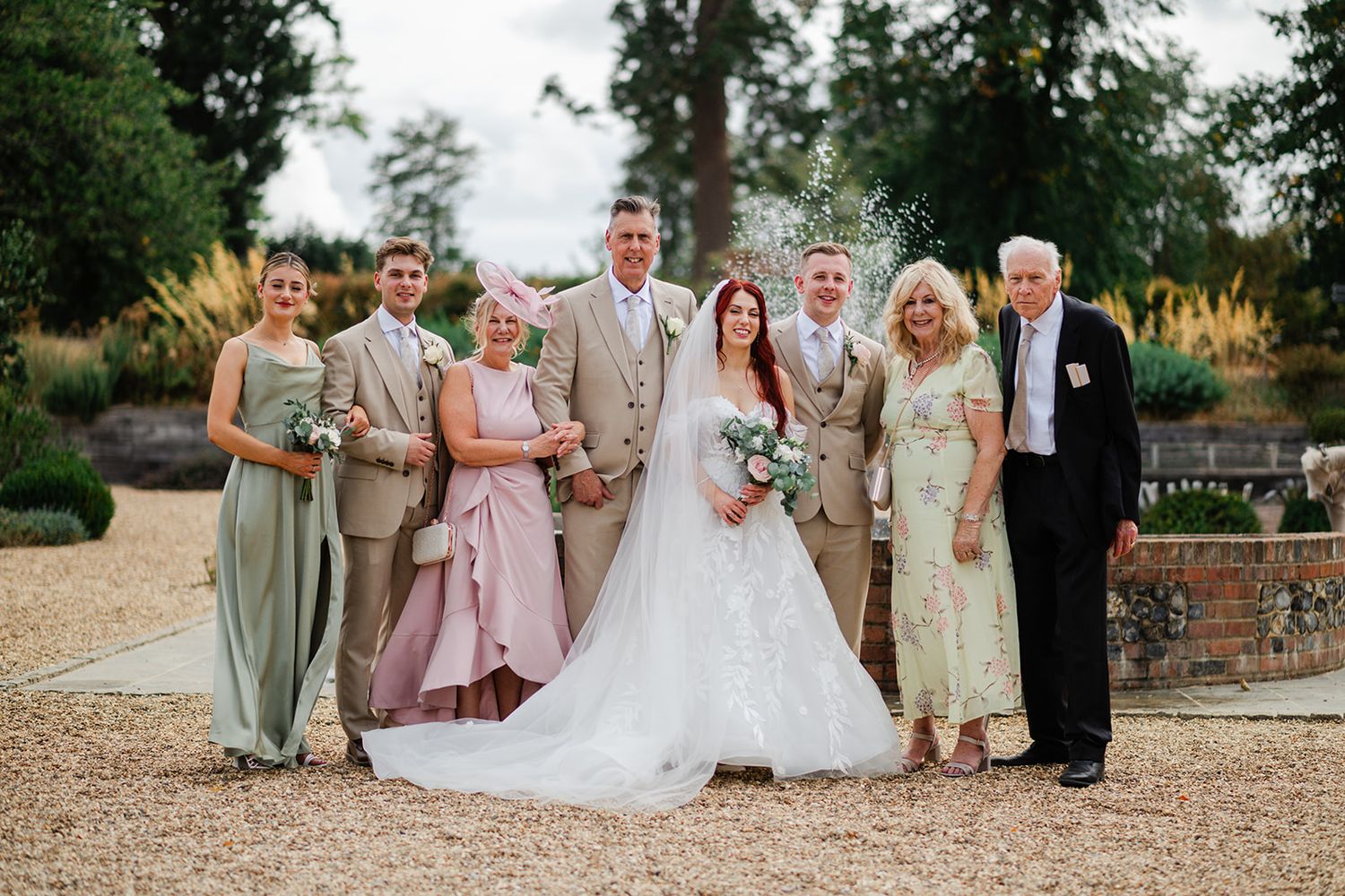 Wedding party poses outdoors, bride in white gown with groom and family in formal attire on gravel path.